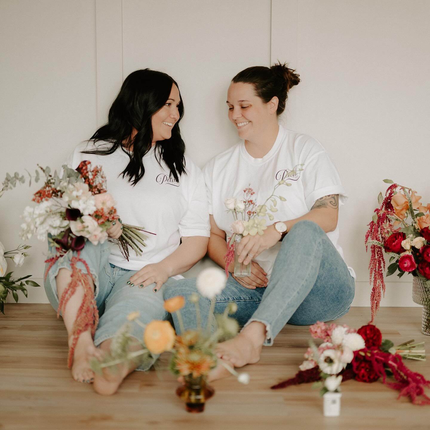 two women in jeans and white shirts sitting on the floor surron=unded by flowers