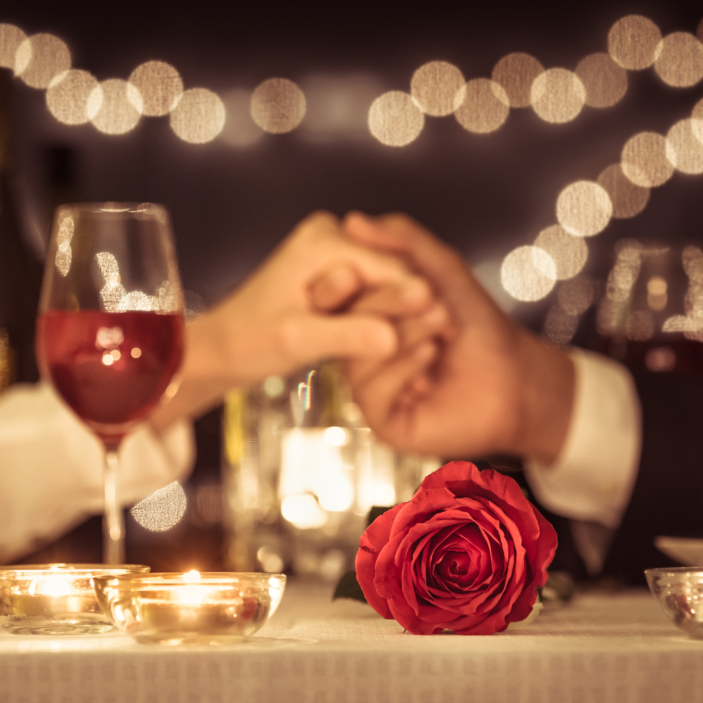 Two adults holding hands at a dinner table with red roses and wine. Valentine's Day inspired