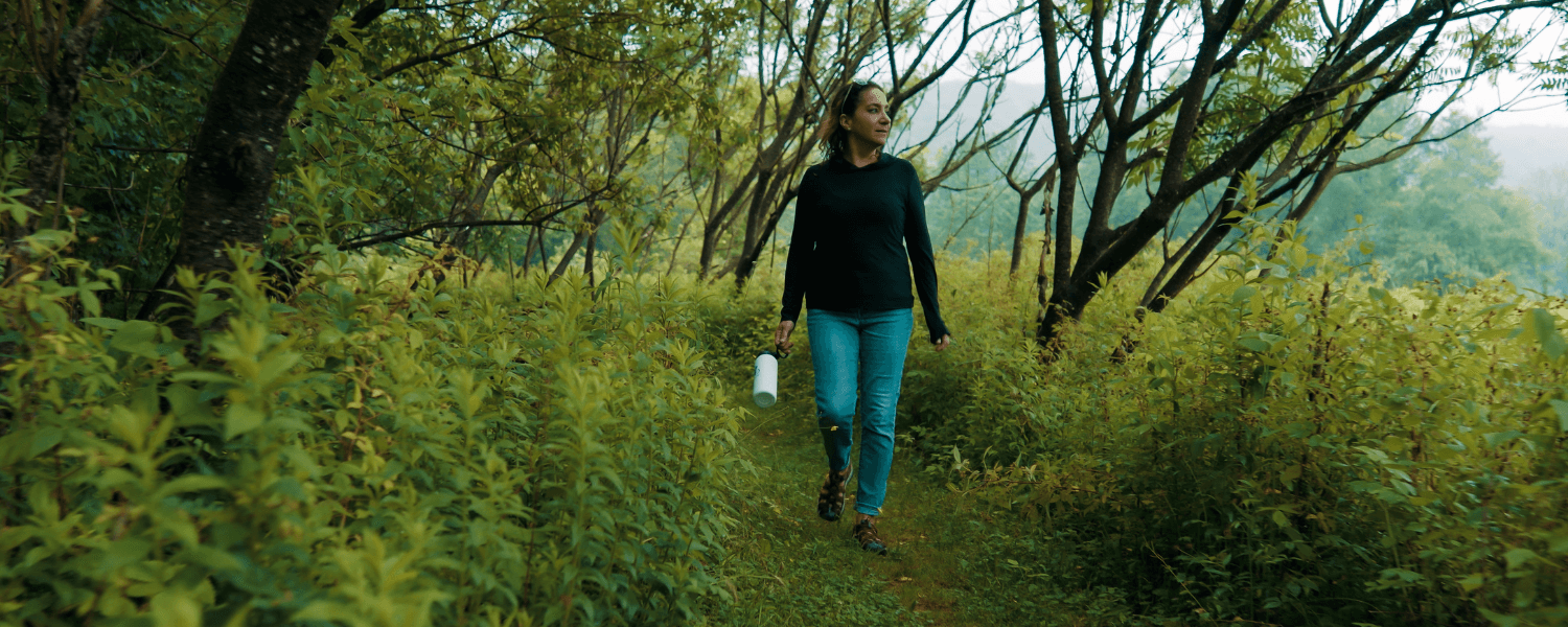 Woman wearing jeans and sweater carrying a water bottle walking in a green wooded area